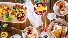 French toasts, chocolatines, cut fruit bowls, fruit salad, scrambled eggs, sunglasses, coffee mug, cinnamon rolls, berry smoothie and ketchup set on a wooden table
