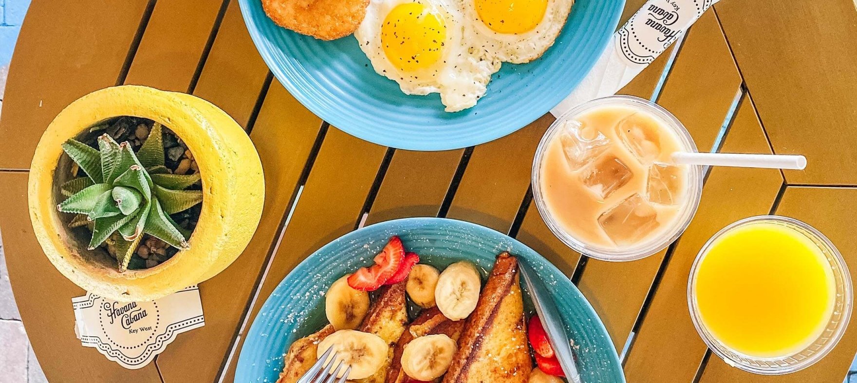 A pair of hands using cutleries on french toasts while another hand is holding up a smoothie next to a egg and bacon breakfast