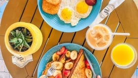 A pair of hands using cutleries on french toasts while another hand is holding up a smoothie next to a egg and bacon breakfast