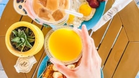 Two hands clinking drinks over a plate of french toast garnished with sliced fruits next to a plate of eggs and bacon.