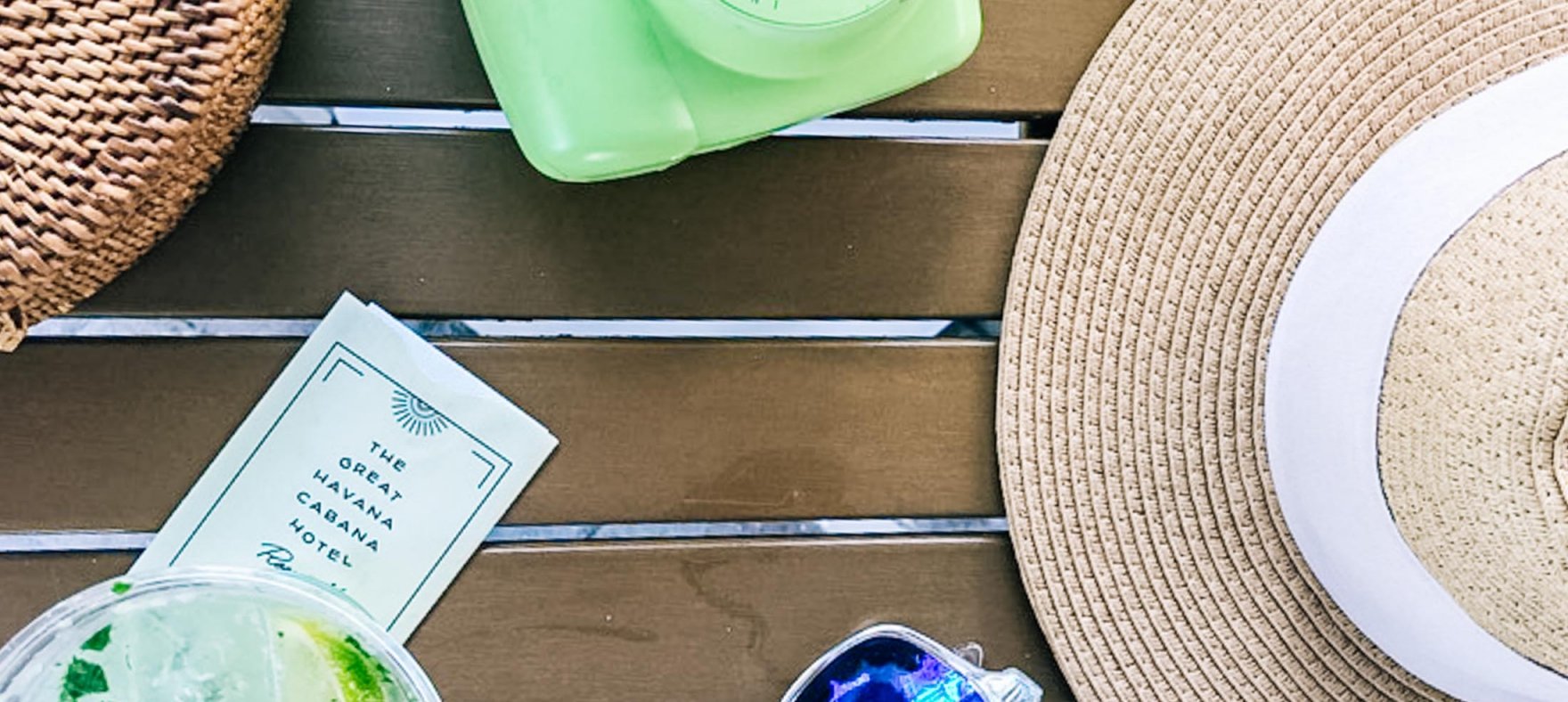 A straw hat next to a pair of sunglasses, a glass of mojito, Havana Cabana collectible card, woven bowl, an instant camera and a smartwatch