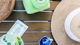 A straw hat next to a pair of sunglasses, a glass of mojito, Havana Cabana collectible card, woven bowl, an instant camera and a smartwatch