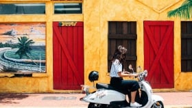 A woman rides a scooter past a yellow house.