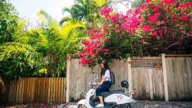 A woman poses on a scooter next to a row of fences and trees.