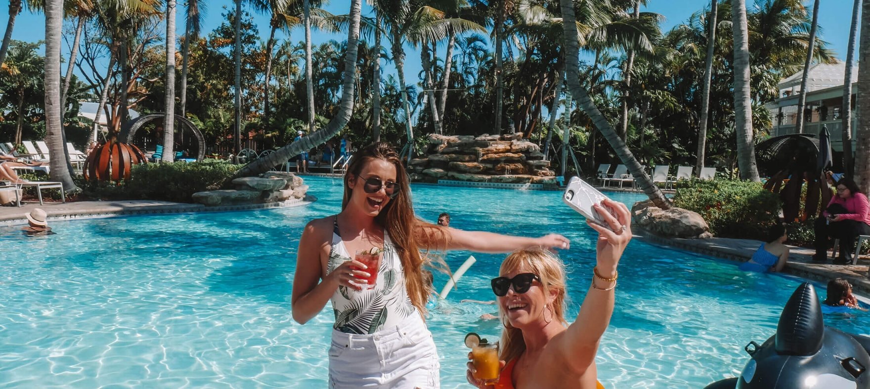 Two women posing for a selfie with drinks in their hands with the outdoor pool in the background