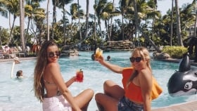 A back shot of two women sitting by the pool and raising their cocktails as they look back