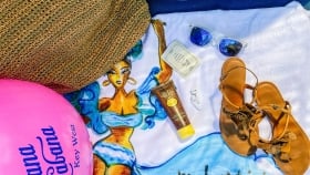 Beach ball, a straw-woven bag placed on a towel next to a sunscreen tube, ear pods, a card, sunglasses and a pair of strappy sandals
