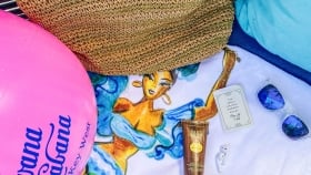 Beach ball, a straw-woven bag placed on a towel next to a sunscreen tube, ear pods, a card, sunglasses
