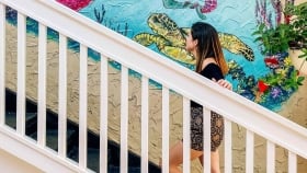 A woman walking up a flight of wooden stairs next to a textured mural of a mermaid next to sea horses and turtles under water