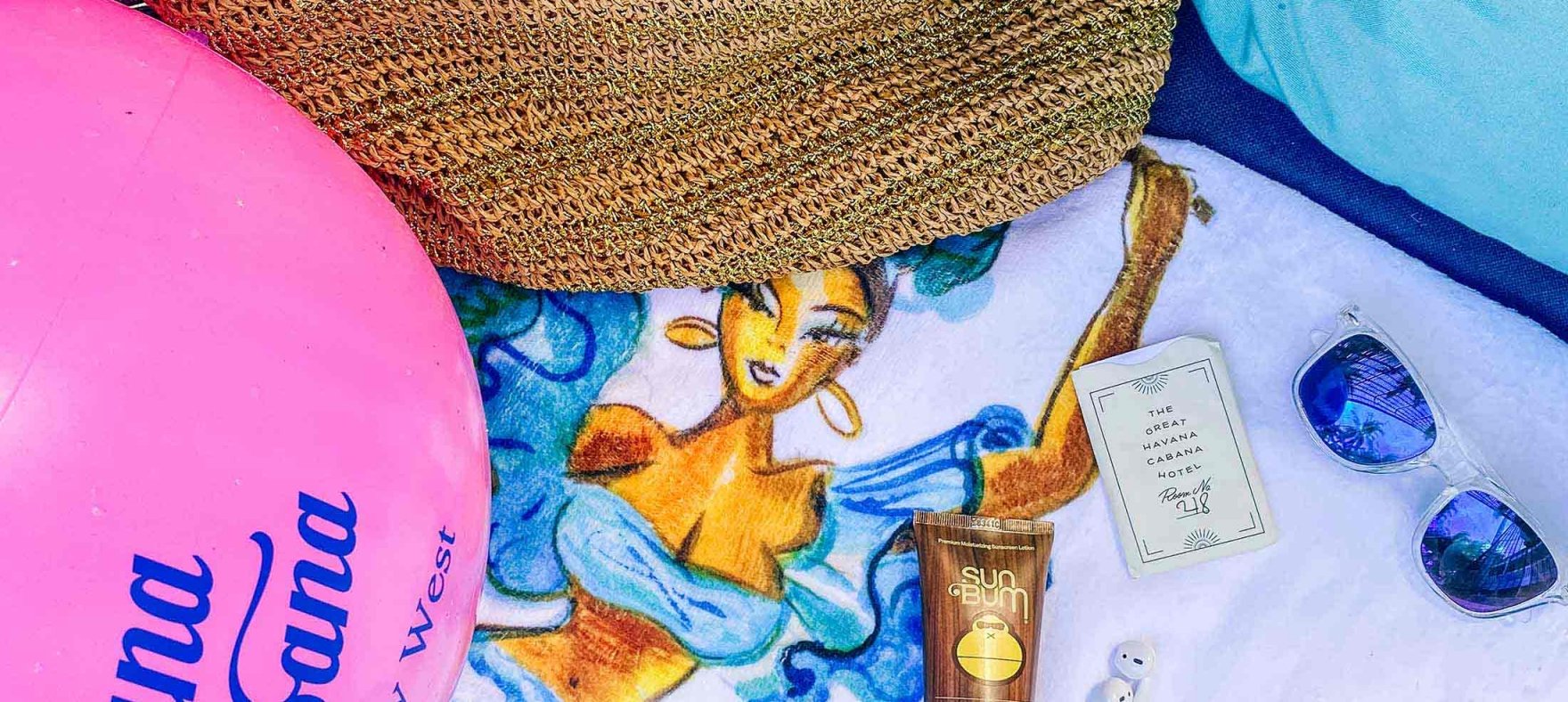 Beach ball next to a woven bag, sunscreen, EarPods, cards and a sunglass placed on a fabric with a painting of a dancer