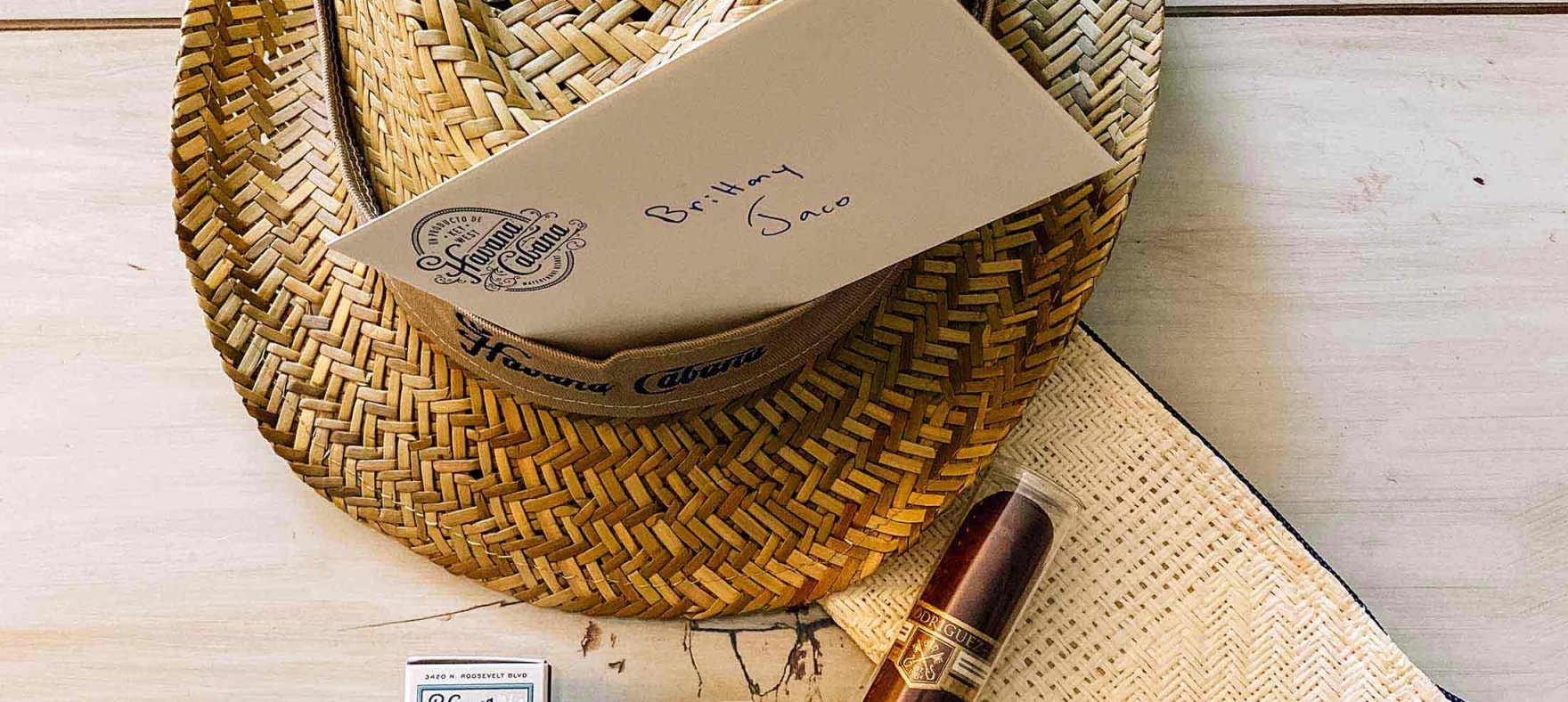 A matchbox, cigar cutter, a Cuban cigar and a HC initialled napkin placed in front of a straw fedora with a letter