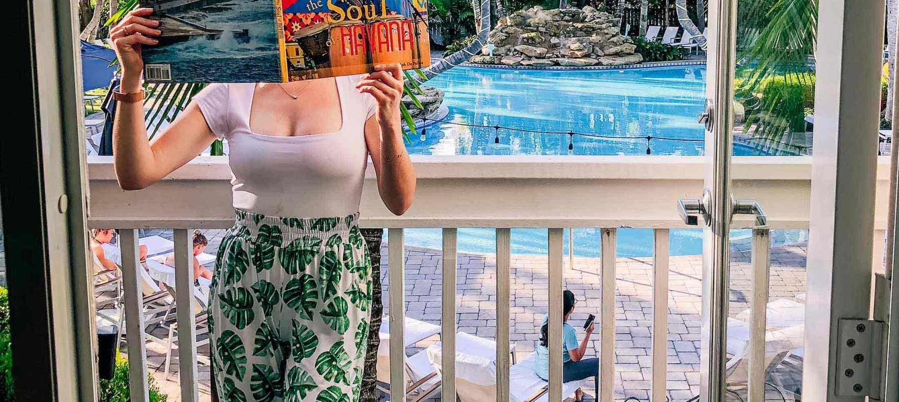 A vertical shot of a woman covering a face with a magazine as she poses in the balcony overlooking the outdoor pool