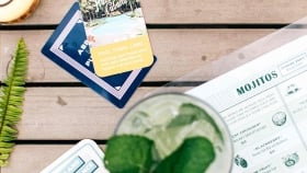 A top shot of a mojito sitting on top of a menu on a wooden table with collectable cards and a glass of Pina Colada.