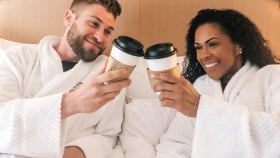 A couple in robes sitting on a bed and clinking their disposable coffee cups