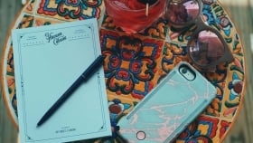 A pen set on a notepad, next to a faced-down cellphone, a pair of sunglasses and a cocktail on a multi-colored table