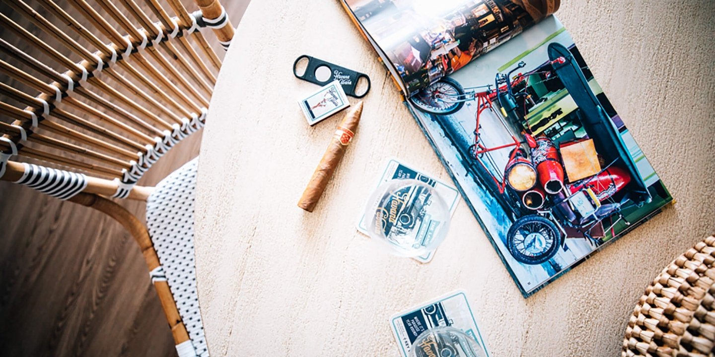 top shot of a cigar, matchbox and a cigar cutter next to wine glasses placed on coasters and a magazine laid out