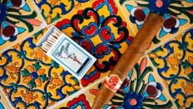 A close up of a Cuban cigar next to a Havana Cabana matchbox placed on a multi-colored background with intricate designs
