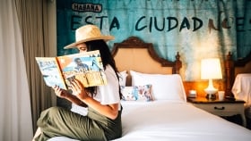 A woman sitting on a bedside, wearing a straw hat and reading a book