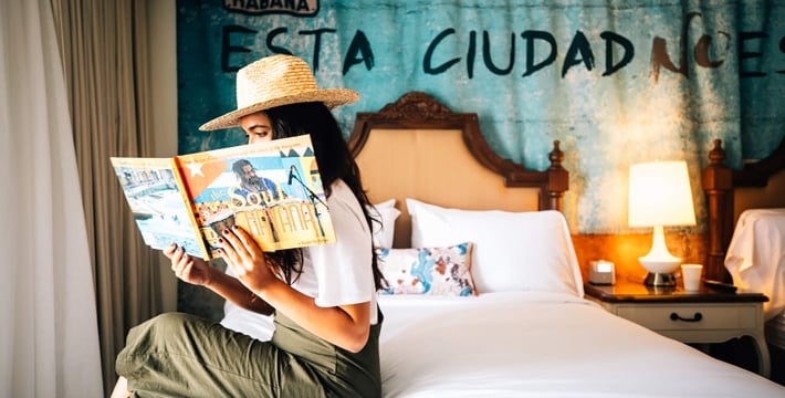 A woman sitting on a bedside, wearing a straw hat and reading a book