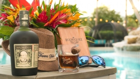 A poolside table with a Diplomatico rum bottle next to a fedora, rum glass with a cigar holder, a sunglass and a wooden placard