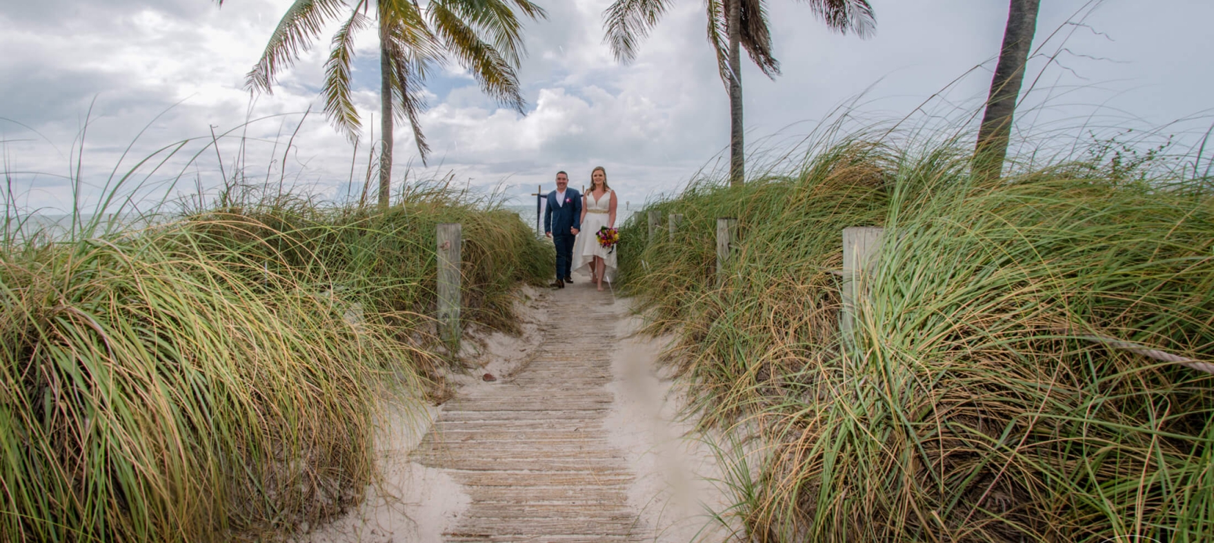 A long shot of the bride and groom standing on a pathway between lush greenery at the beach