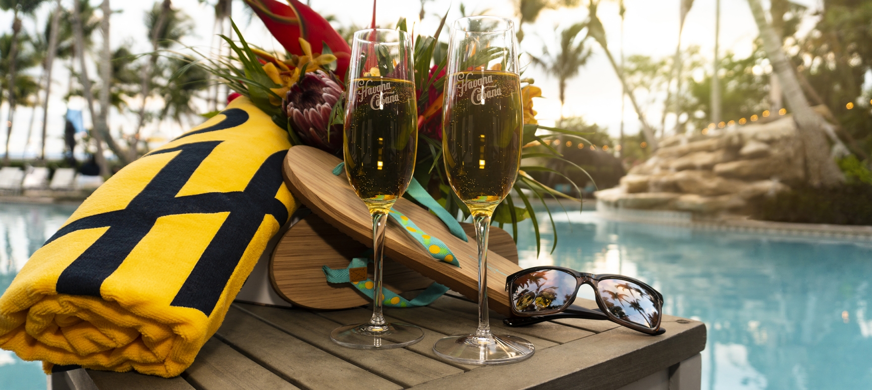 A close-up of a pair of champagne flutes next to flip-flops, rolled towel, sunglasses and a bouquet of bright flowers.