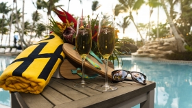 A close-up of a pair of champagne flutes next to flip-flops, rolled towel, sunglasses and a bouquet of bright flowers.