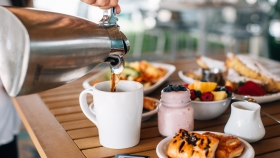 A hand pouring coffee into a mug from a steel coffee pot next to a pair of chocolatines, a berry smoothie, a bowl of cut fruits and more