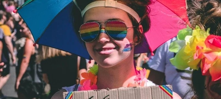 Woman wearing a colorful umbrella hat with rainbow-tinted sunglasses and colours smeared on her right cheek